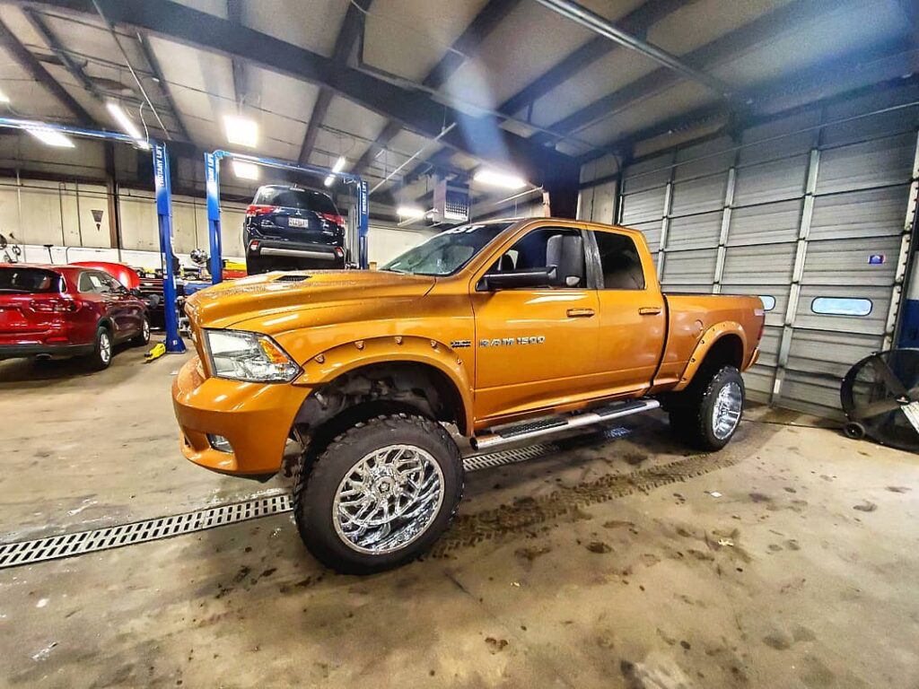 A lifted orange Ram 1500 on polished wheels inside the Apple Off Road shop, under the overhead lights.